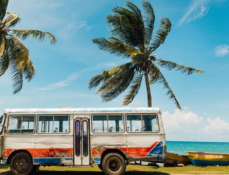An einer Strandpromenade steht ein alter, rustikaler bunter Bus. Daneben und dahinter Kokospalmen.