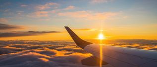 Blick aus einem Flugzeugfenster auf den Flügel, über den Wolken und die untergehende Sonne, die das Bild in gold-gelbes Licht taucht.