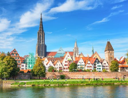 An einem Flussufer mit anliegendem Grünstreifen stehen süße, kleine Fachwerkhäuser und eine schmale, spitze Kirche ragt in den blauen Himmel.