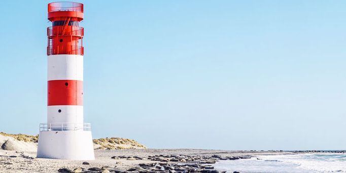 Am Strand steht ein weiß-roter Leuchtturm und dahinter beginnen die Dünen.
