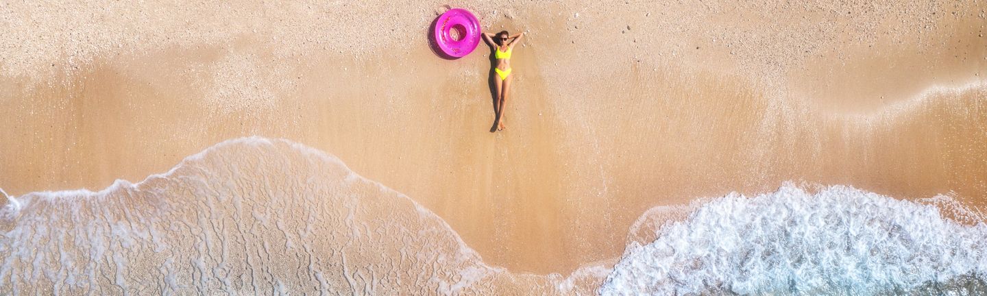Eine Frau im gelben Bikini liegt entspannt am Strand. Neben ihr befindet sich ein pinker Gummi Donut.