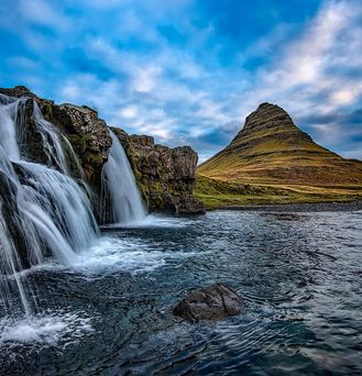 Ein spitzer, Zwergenhut-artiger Berg ragt aus einer grünen Landschaft empor. Im Vordergrund ein Fluss, See oder Meeresarm, in welchen von einem Felsabhang Wasser fließt.