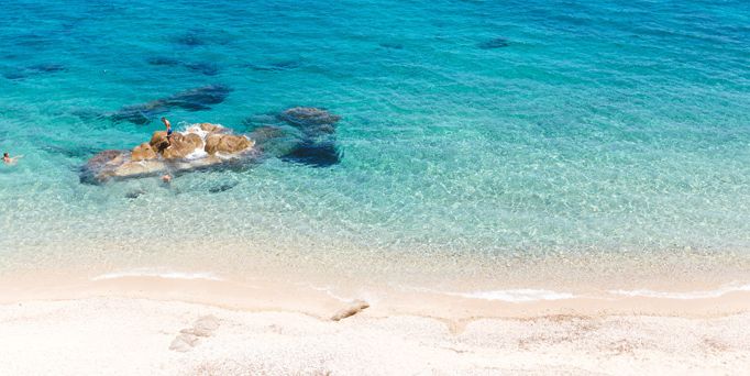 Kristallklares, türkises Wasser an einem weißen Sandstrand. Im Wasser ein kleiner Felsen.