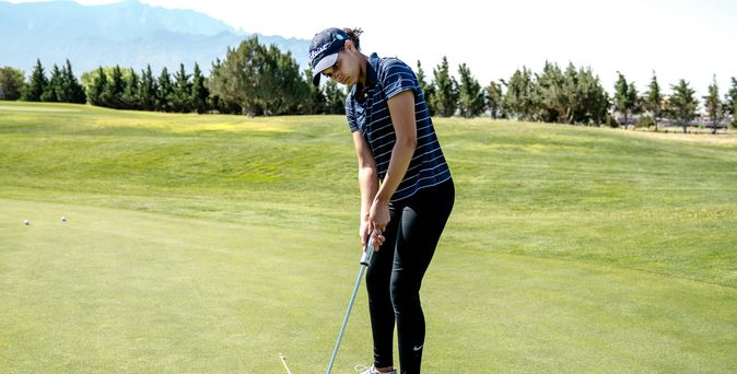 Eine Person in Golfkleidung und Cap steht auf einem grünen Golfplatz und hat gerade gegen den Golfball geschlagen, welcher kurz vor dem Loch liegt.