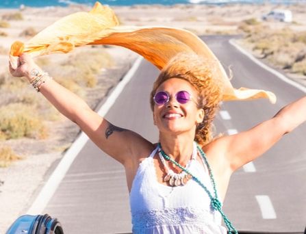Eine sommerlich, hippimäßig gekleidete Frau sitzt in einem Cabrio mit offenem Dach. Sie steht im Auto und reist die Arme lachend nach oben und ein orangenes Tuch weht im Wind. Im Hintergrund das wilde Meer.