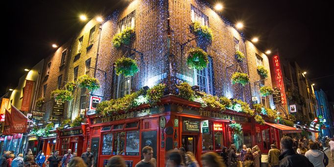 Ein traditionell irisches Backstein-Eckhaus mit roten Fensterläden und Irish-Pubs im dunkel. Davor tummeln sich Menschenmassen durch das nächliche Dublin.