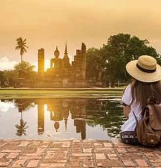 Eine junge Frau mit Sonnenhut und Rucksack sitzt vor einem buddhistischem Tempel.