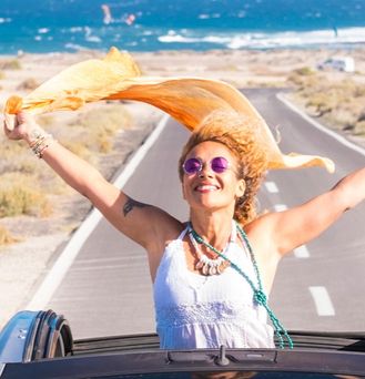 Eine sommerlich, hippimäßig gekleidete Frau sitzt in einem Cabrio mit offenem Dach. Sie steht im Auto und reist die Arme lachend nach oben und ein orangenes Tuch weht im Wind. Im Hintergrund das wilde Meer.