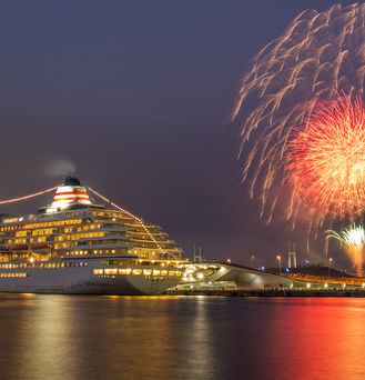 Ein beleuchtetes Kreuzfahrtschiff befindet sich an einem Hafen. Es ist dunkel und im Hintergrund leichtet das Feuerwerk.