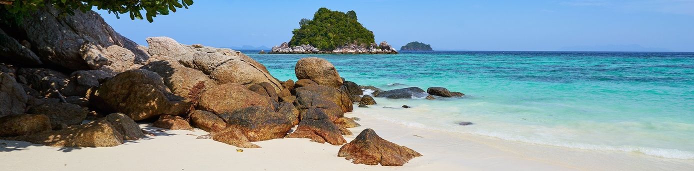 Ein feinsandiger, weißer Strand mit großen Steinen, welche ins türkisblaue, klare Meer ragen. Im Hintergrund eine kleine Insel.