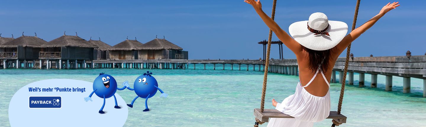 Malediven: Eine Frau in weißem Kleid und mit Sonnenhut schaukelt auf einer Holzschaukel und reißt die Arme in die Luft. Sie blickt auf den glasklaren türkisen Ozean mit einem Holzsteg und Wasserbungalows.
