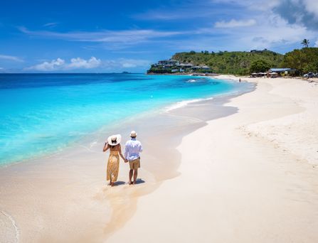 Ein Paar in Sommerkleidung läuft Hand in Hand an einem weißen Sandstrand entlang. Das Wasser ist türkisblau und im Hintergrund tropischer Wald und einzelne Häuser.