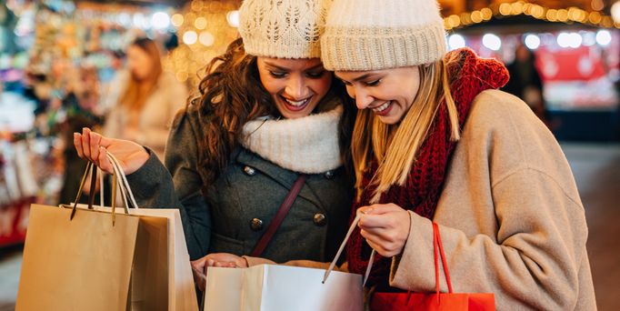 Zwei winterlich gekleidete Frauen schauen begeistert in ihre Shoppingtaschen. Im Hintergrund erahnt man Weihnachtsmarktbeleuchtung