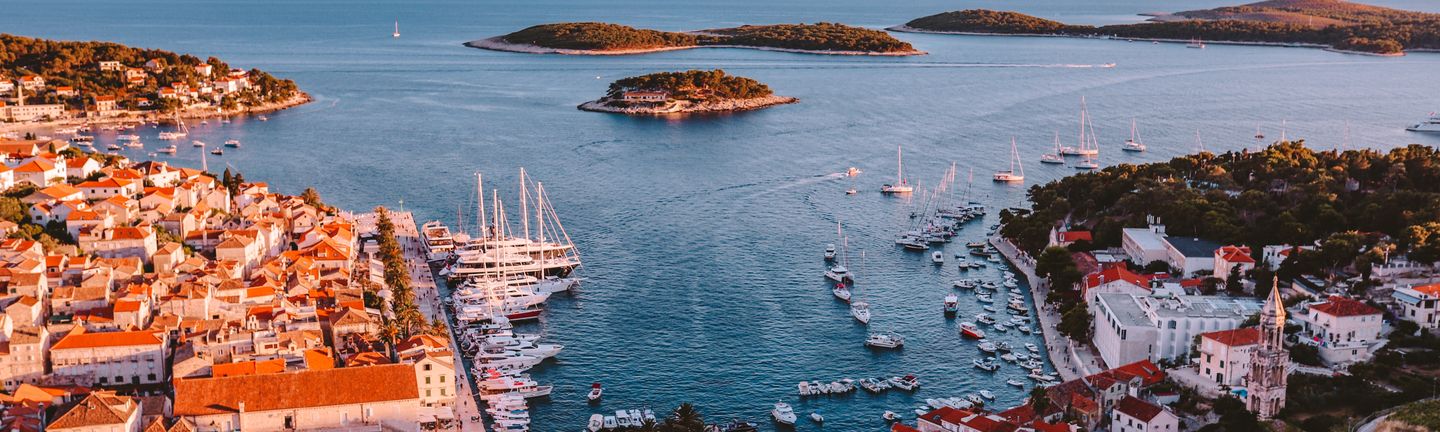 Eine Luftaufnahme einer belebten Bucht mit Hafen mit vielen Seegelbooten, einem malerischen Dorf mit orangenen Dächern und 2 bis 3 Inseln.