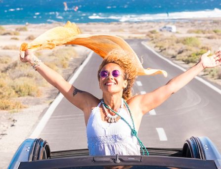 Eine sommerlich, hippimäßig gekleidete Frau sitzt in einem Cabrio mit offenem Dach. Sie steht im Auto und reist die Arme lachend nach oben und ein orangenes Tuch weht im Wind. Im Hintergrund das wilde Meer.