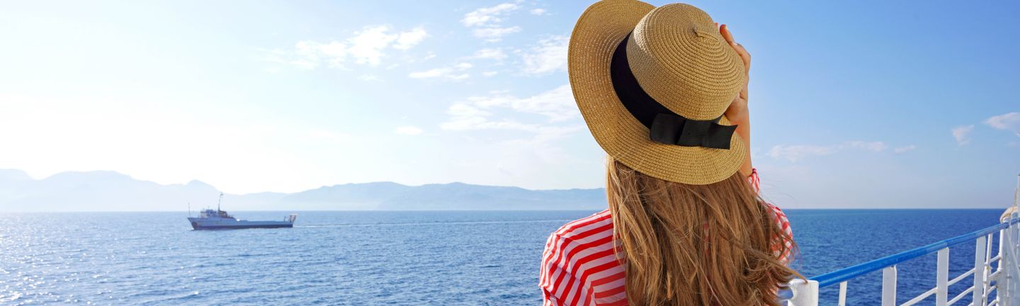 Eine blonde Frau mit Sonnenhut steht an der Rehling eines Kreuzfahrtschiffes und blickt aufs Meer.