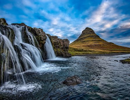 Ein spitzer, Zwergenhut-artiger Berg ragt aus einer grünen Landschaft empor. Im Vordergrund ein Fluss, See oder Meeresarm, in welchen von einem Felsabhang Wasser fließt.