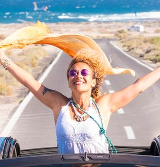 Eine sommerlich, hippimäßig gekleidete Frau sitzt in einem Cabrio mit offenem Dach. Sie steht im Auto und reist die Arme lachend nach oben und ein orangenes Tuch weht im Wind. Im Hintergrund das wilde Meer.